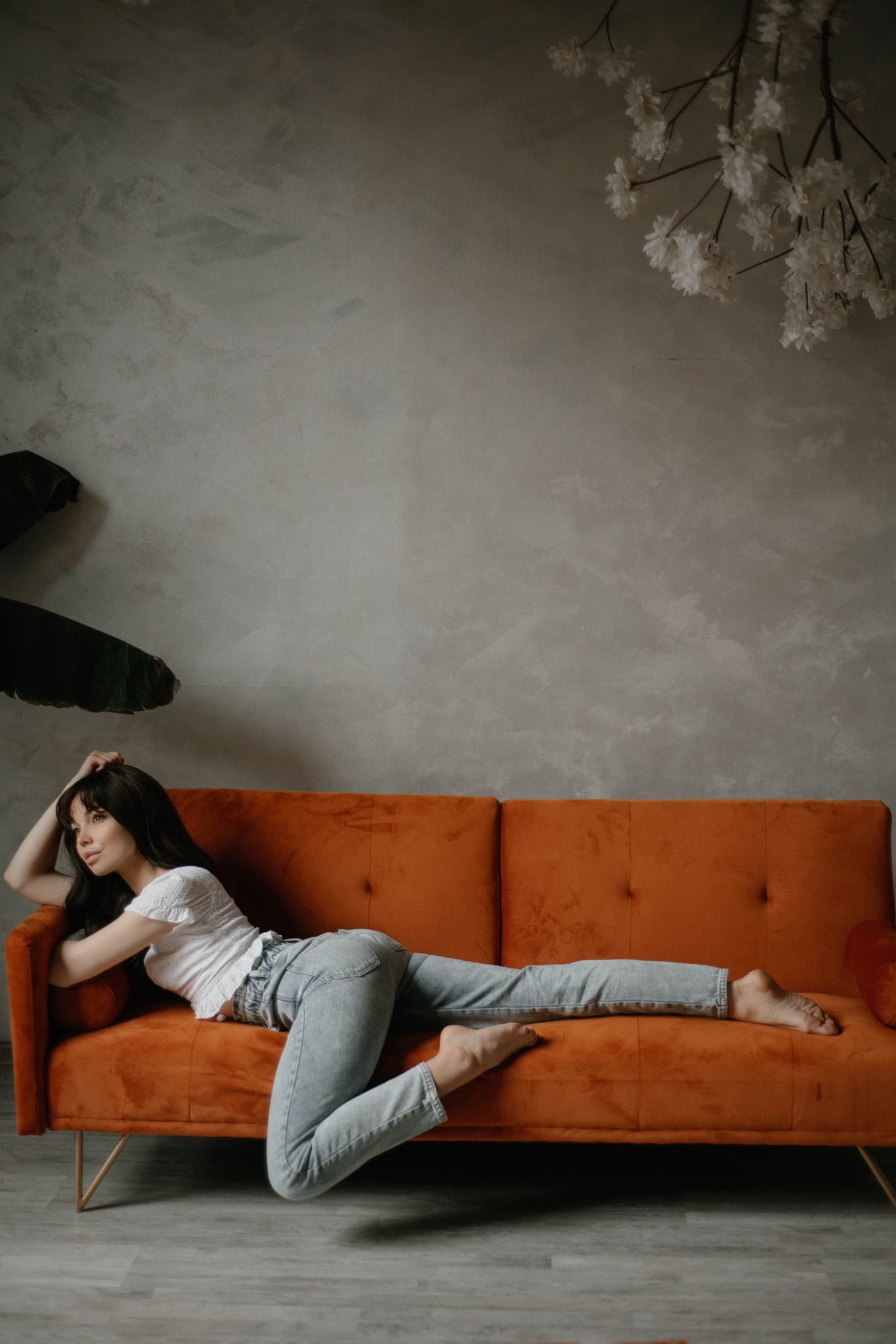 Kobieta w jasnych jeansach i białej bluzce leży na pomarańczowej welurowej sofie na tle szarej ściany z dekoracją kwiatową