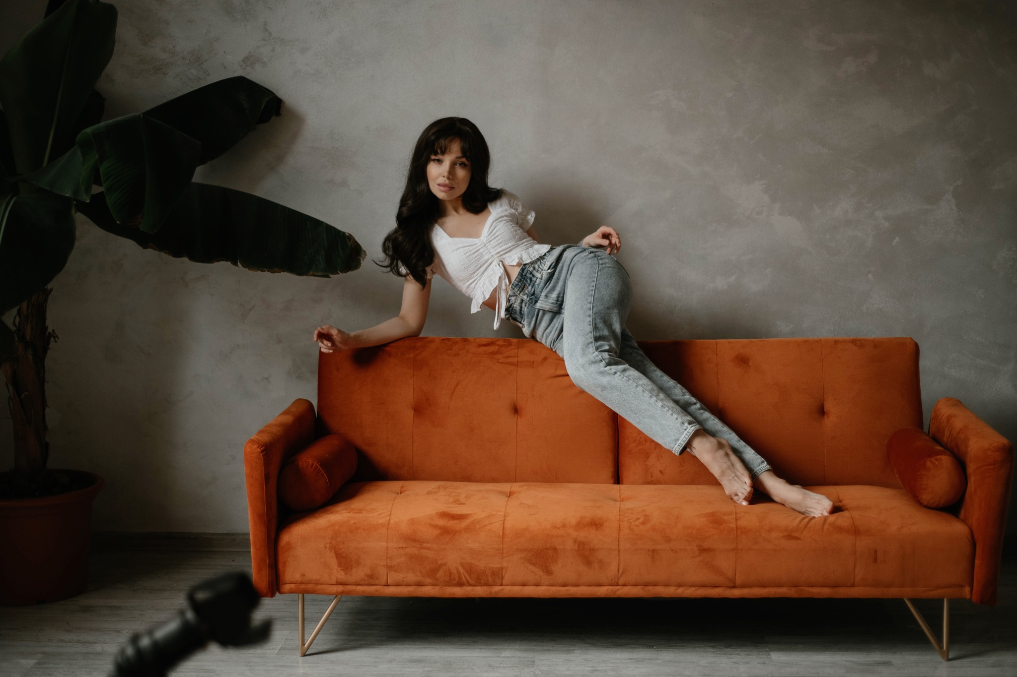 Młoda kobieta w jasnych jeansach i białej bluzce pozuje na pomarańczowej welurowej sofie w studio fotograficznym, obok dużej rośliny doniczkowej.
