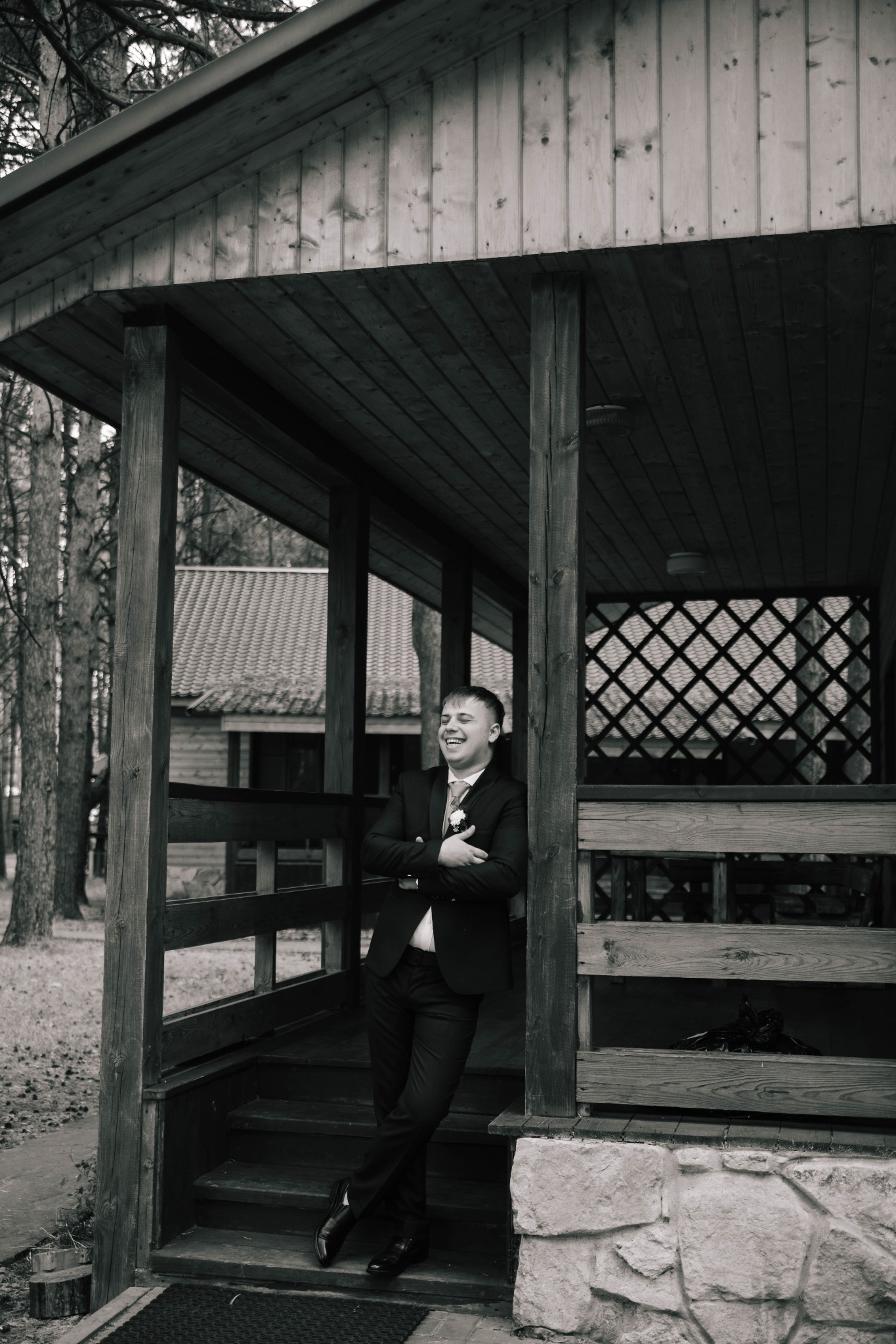 Uśmiechnięty mężczyzna w garniturze, oparty o drewnianą werandę z ażurową ścianą, w tle widoczny budynek z dwuspadowym dachem.