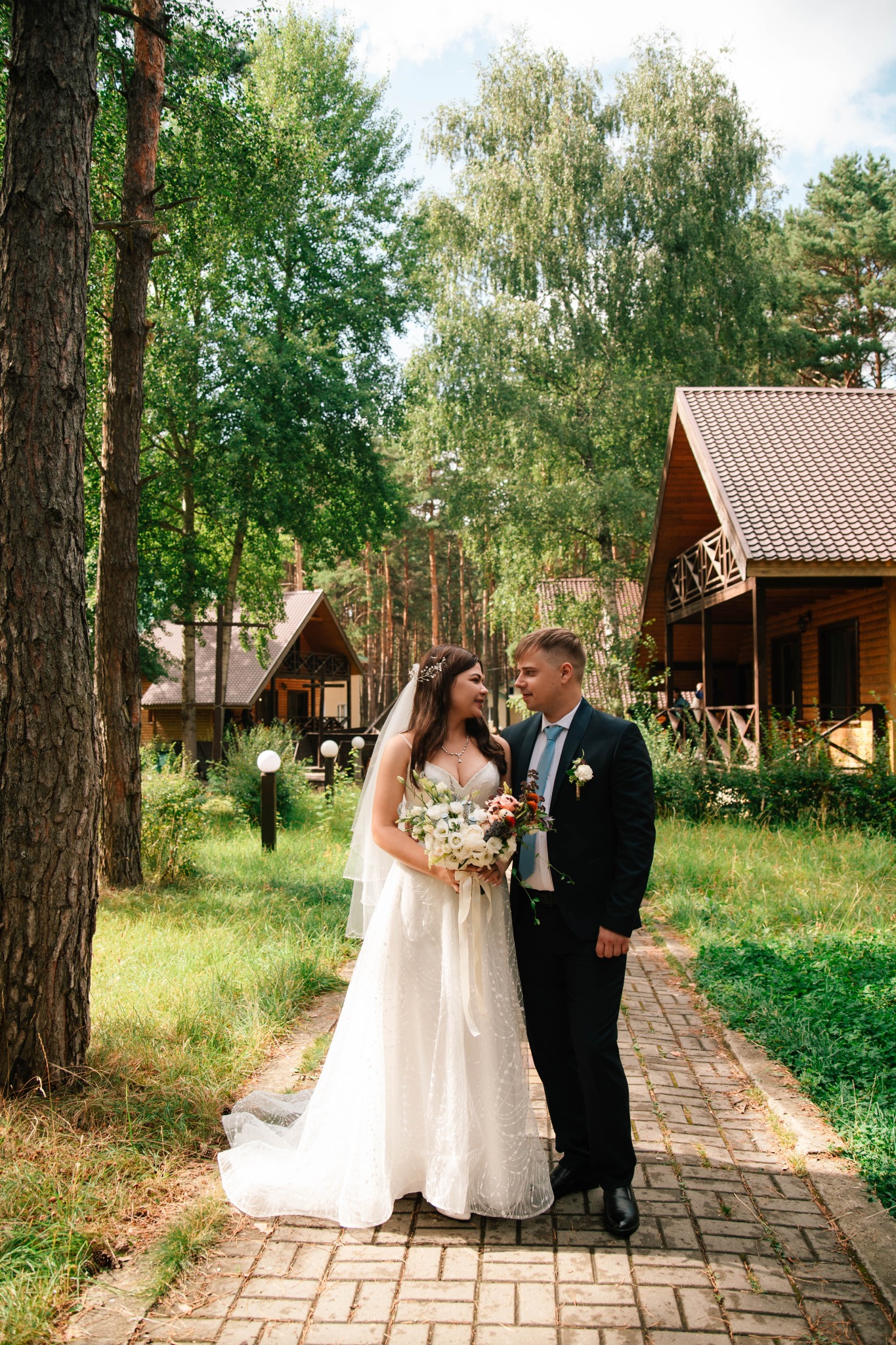 Para młoda w plenerze, panna młoda w białej sukni z bukietem kwiatów, pan młody w czarnym garniturze i błękitnym krawacie, na tle drewnianych domków i zieleni.