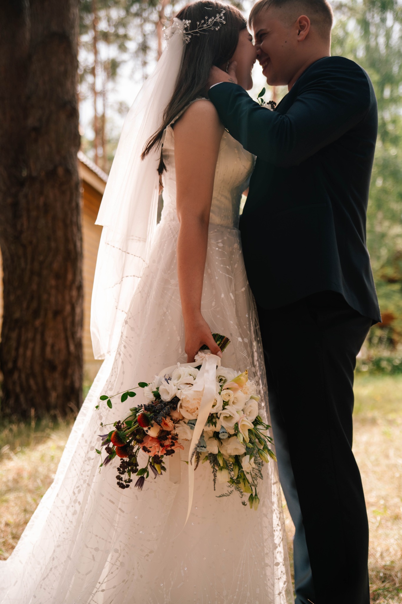 Panna młoda w białej sukni trzyma bukiet ślubny, zbliżenie z panem młodym w ciemnym garniturze w plenerze.