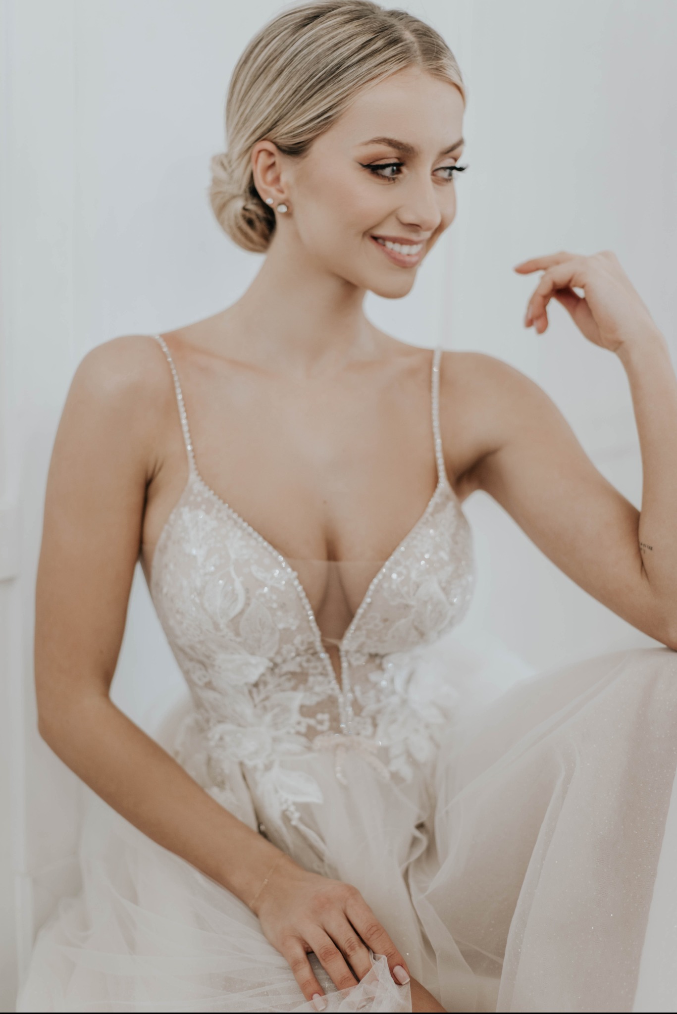 Uśmiechnięta panna młoda w sukni ślubnej z głębokim dekoltem, siedząca na białym tle, z upiętymi blond włosami i delikatnym makijażem.