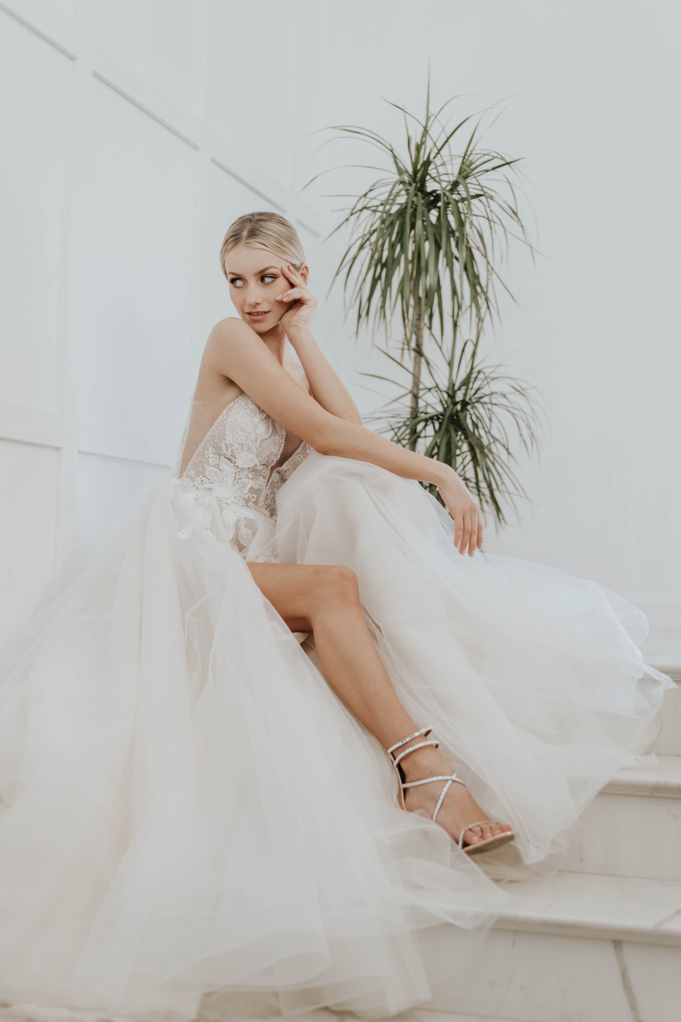 Panna młoda w białej sukni ślubnej z koronkowym gorsetem i tiulowym dołem, siedząca na marmurowych schodach, w tle biała ściana i zielona roślina doniczkowa.