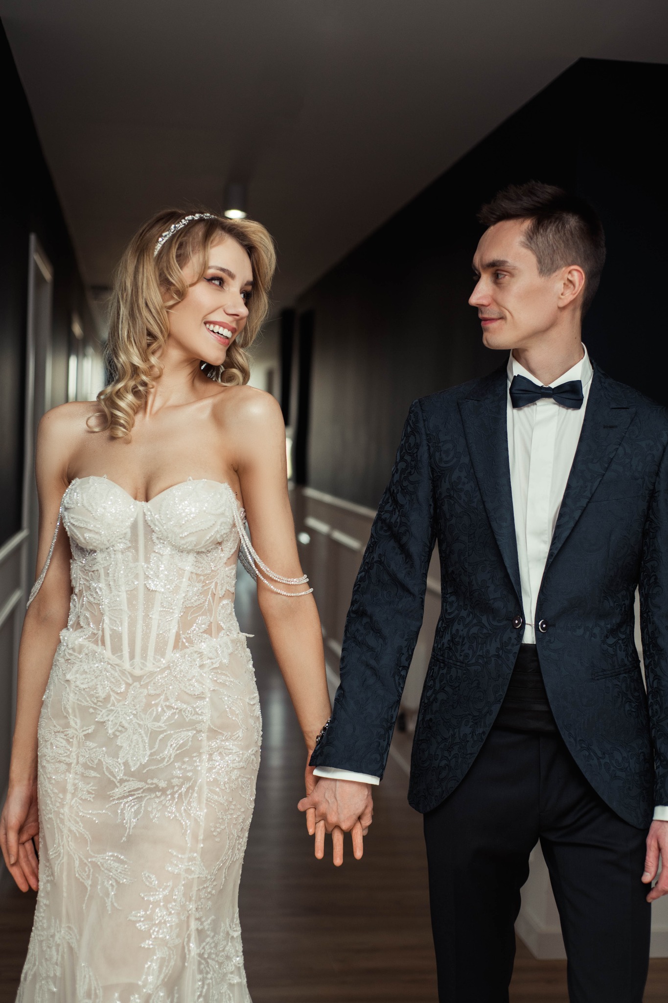 Uśmiechnięta panna młoda w koronkowej sukni bez ramiączek i diademie spogląda na pana młodego w granatowej marynarce w kwiatowy wzór i muszce, trzymając się za ręce w nowoczesnym wnętrzu.