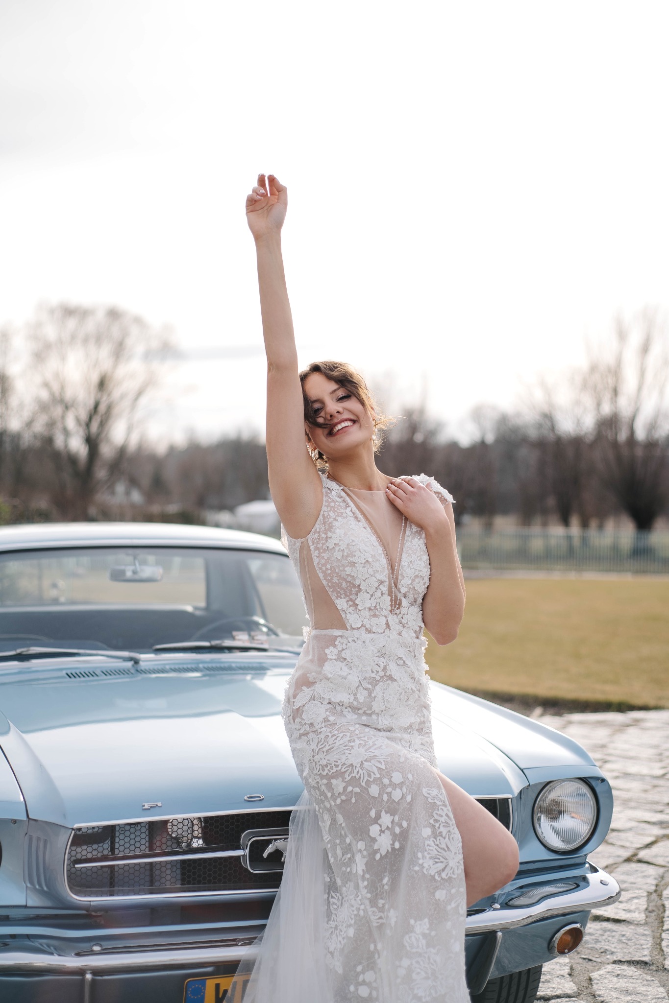 Uśmiechnięta panna młoda w koronkowej sukni, pozująca z uniesioną ręką na masce błękitnego Forda Mustanga z lat 60.