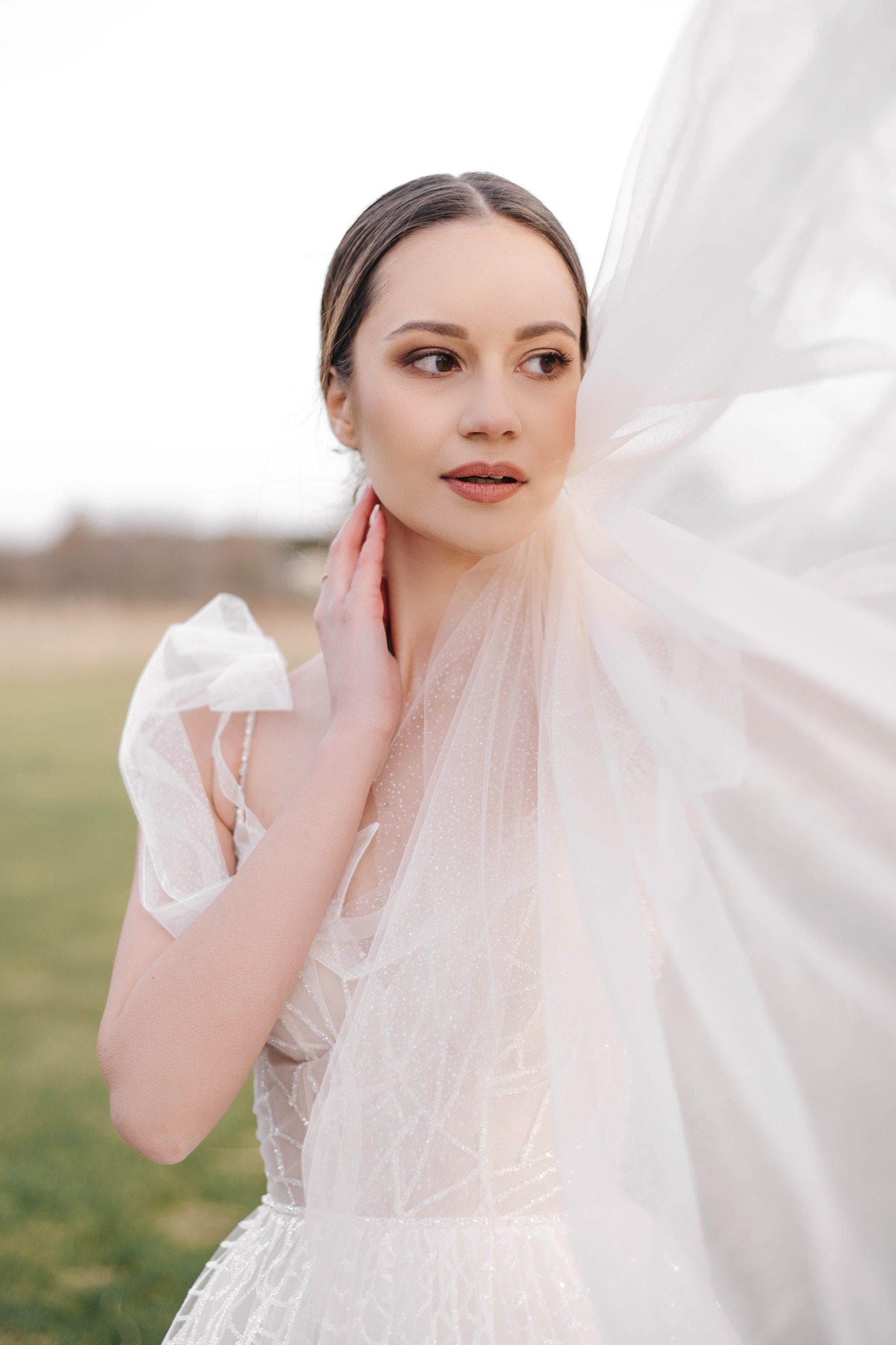 Portret kobiety w zwiewnej, białej sukni ślubnej z delikatnym makijażem, trzymającej dłoń na szyi, z fragmentem welonu powiewającego na wietrze, na rozmytym tle łąki.