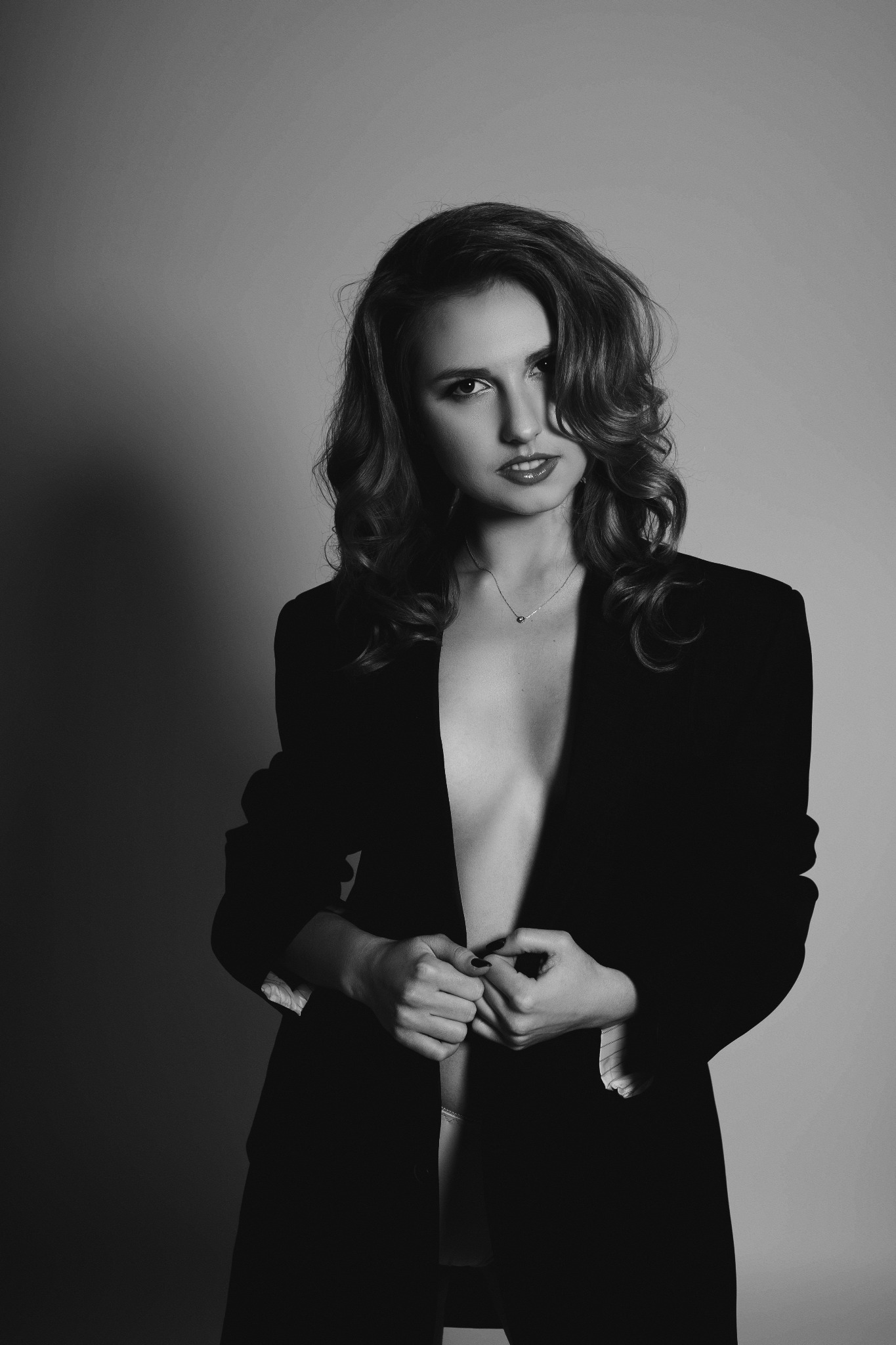 Portret kobiety w czarno-białej tonacji, ubrana w rozpiętą marynarkę, z delikatną biżuterią na szyi, patrząca w obiektyw z wyrazistym makijażem ust.