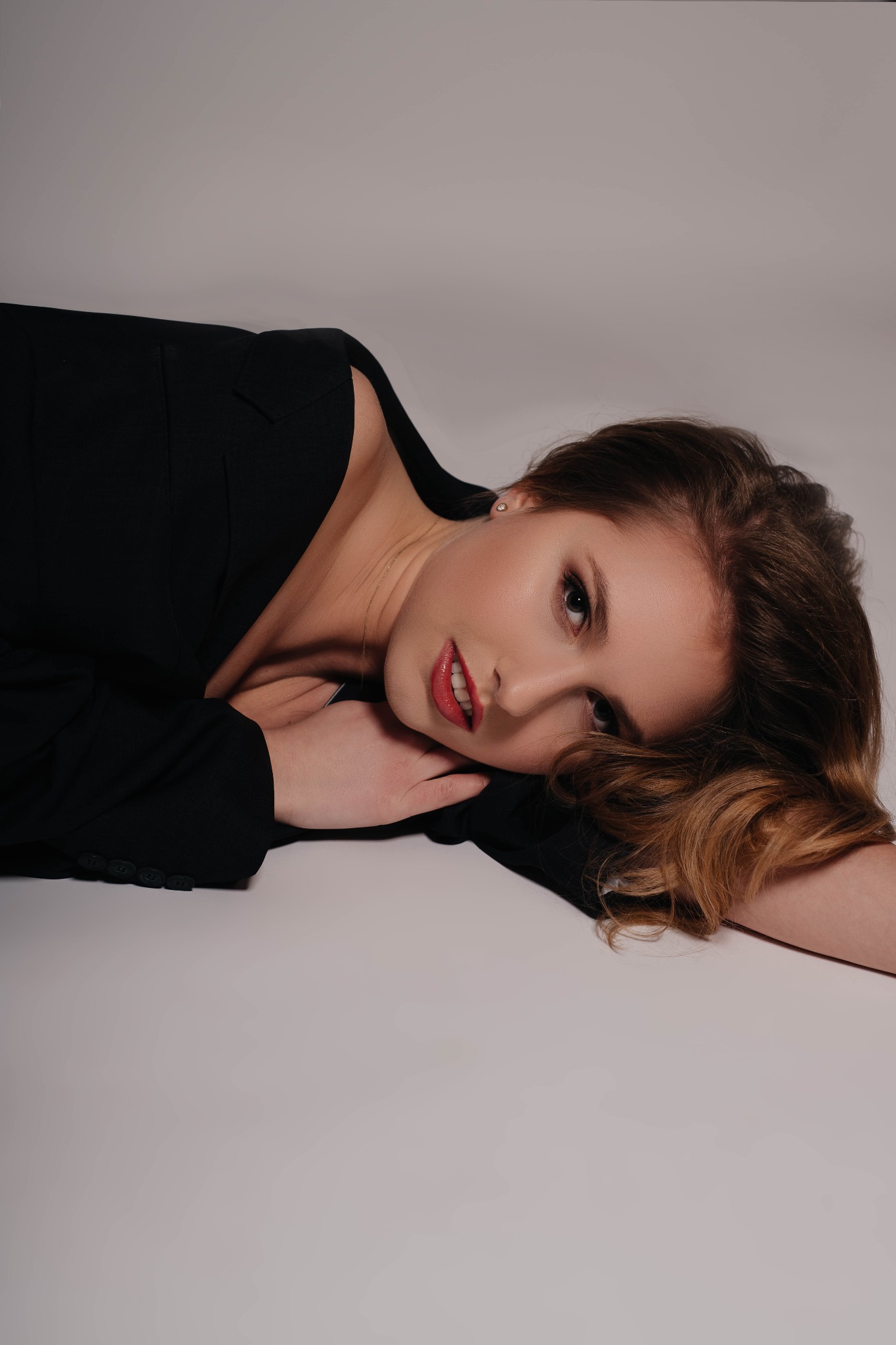 Portret kobiety w czarnej marynarce, leżącej na jasnym tle, z delikatnym makijażem i rozpuszczonymi włosami, spoglądającej w obiektyw.
