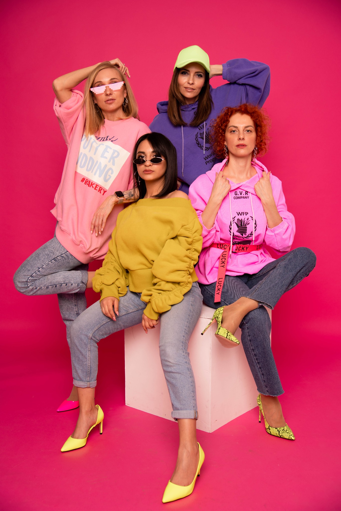 Cztery kobiety w modnych ubraniach pozują na różowym tle, prezentując różnorodne stylizacje i odważne kolory, w tym neonowe szpilki i bluzy z nadrukami.