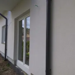 Fragment elewacji domu jednorodzinnego z widoczną strukturą tynku, rynną spustową i oknem z odbiciem zieleni. U podstawy ściany widoczna czarna folia kubełkowa. Na ścianie widoczne ślady po mocowaniu.
