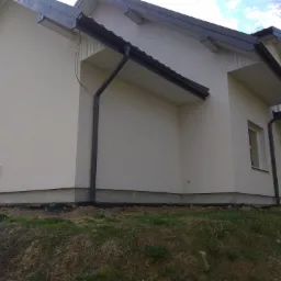 Jasny dom jednorodzinny z ocieploną elewacją i czarnymi rynnami, widoczne fragmenty izolacji fundamentów przy gruncie, trawa i drzewa w tle.