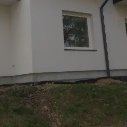 Fragment ocieplonej elewacji budynku z widoczną hydroizolacją fundamentów i oknem z odbiciem drzewa.