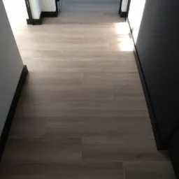 Beżowe płytki podłogowe imitujące drewno w korytarzu z czarnymi listwami przypodłogowymi i ościeżnicami.