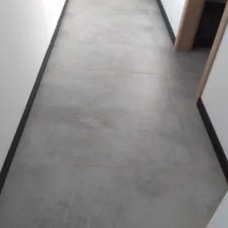 Szare, matowe płytki podłogowe ułożone w korytarzu z białymi ścianami i ciemną listwą przypodłogową; widoczny fragment ościeżnicy drewnianej drzwi.