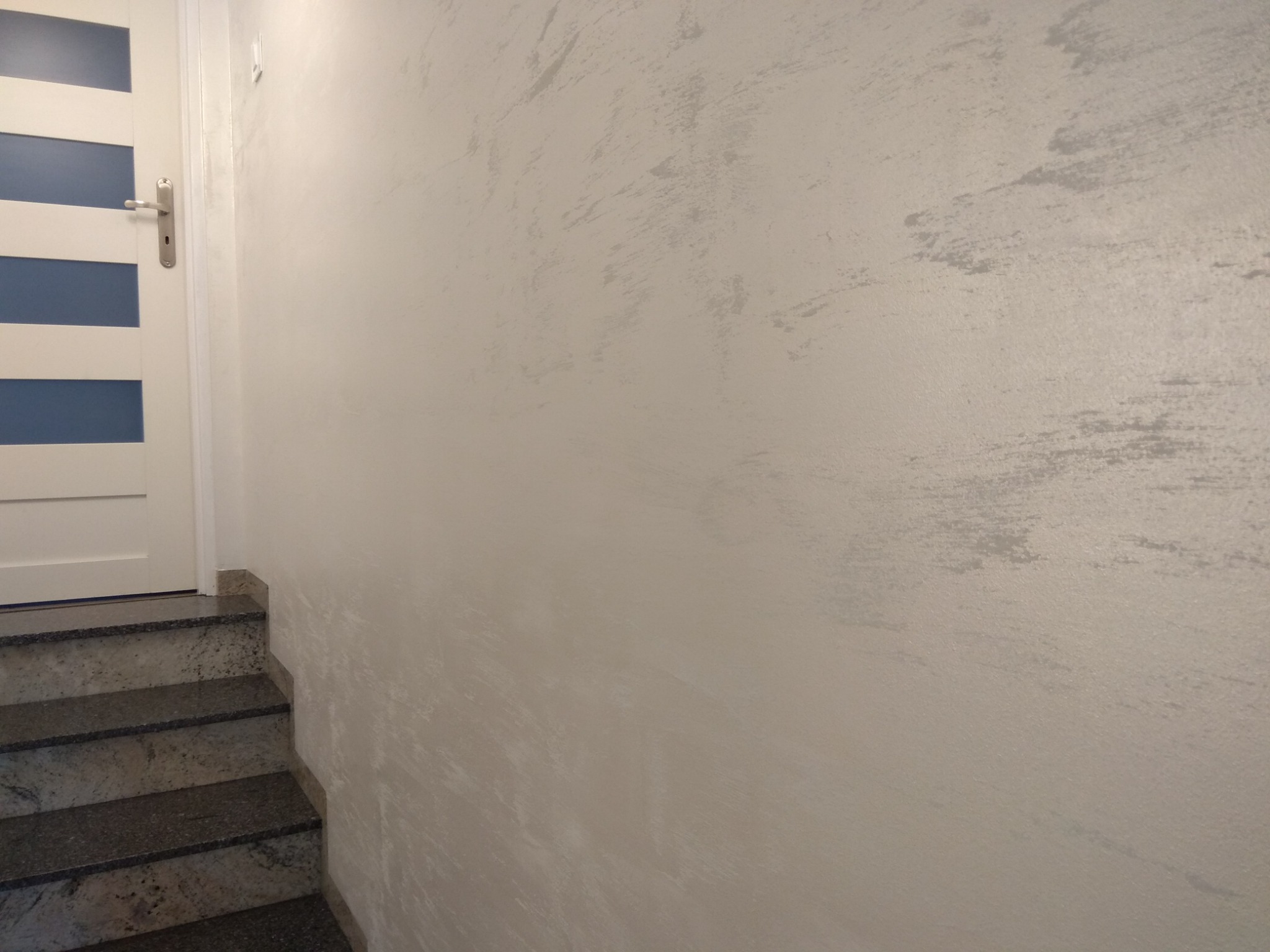 Dekoracyjna ściana z efektem przetarć w odcieniach szarości, białe drzwi z przeszkleniami i granitowe schody.