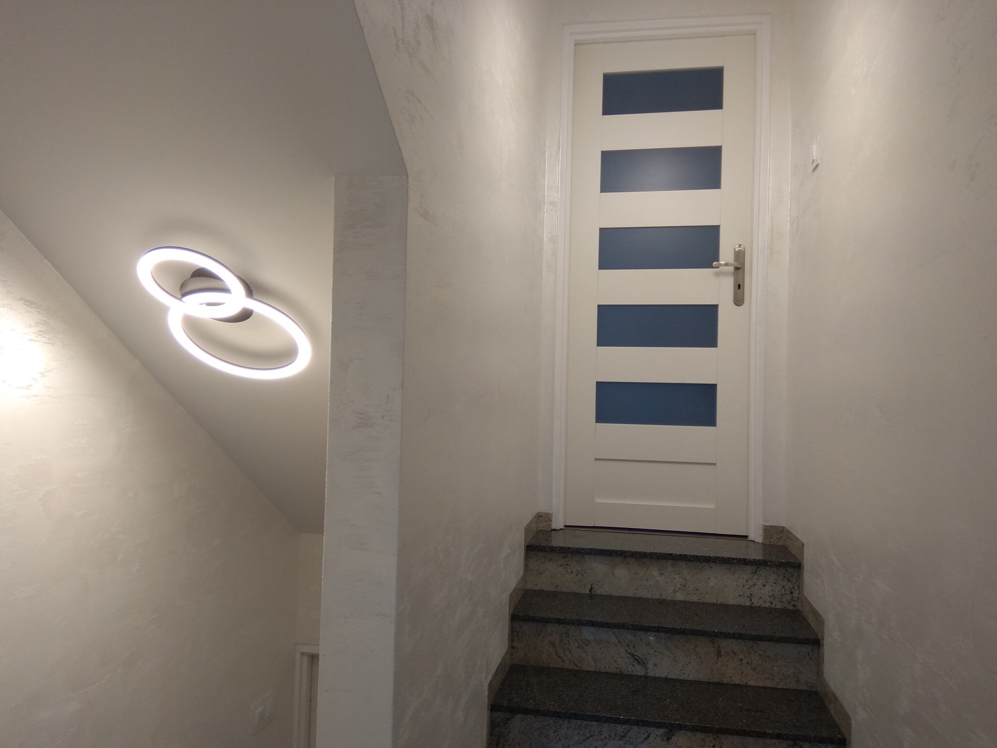 Klatka schodowa z granitowymi schodami, białą ścianą o fakturze trawertynu, nowoczesnym oświetleniem sufitowym w kształcie dwóch połączonych okręgów i białą, przeszkloną w pięciu miejscach...