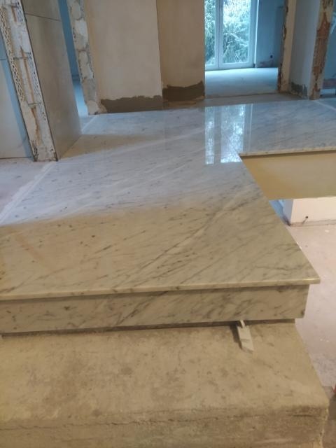 Ułożona marmurowa posadzka w trakcie prac wykończeniowych, widoczne surowe ściany i przygotowane do położenia płytki, okno w tle.