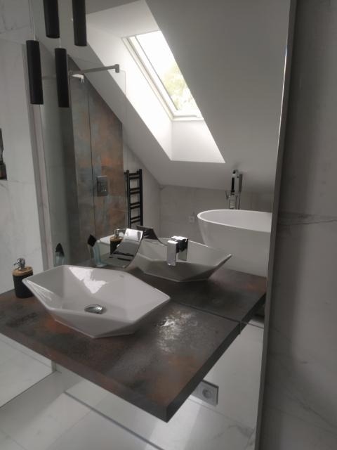 Nowoczesna łazienka z dwiema umywalkami nablatowymi o geometrycznym kształcie, umieszczonymi na blacie imitującym rdzę. W tle widoczna wanna wolnostojąca i okno dachowe.