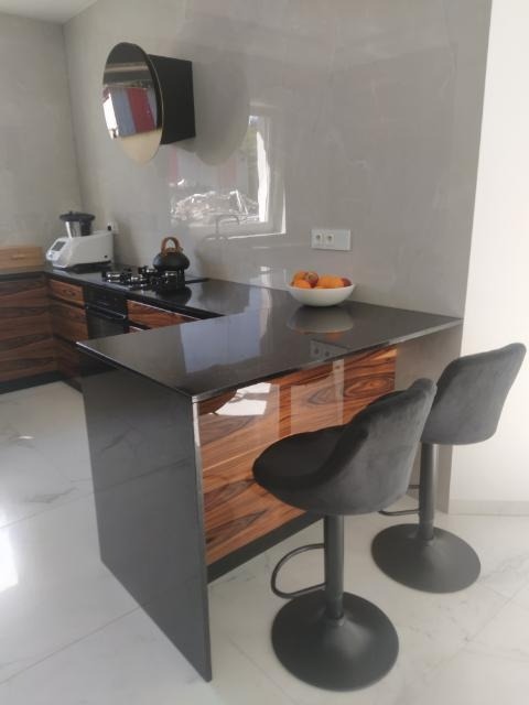 Nowoczesna kuchnia z wyspą wykończoną drewnem, czarne, welurowe hokery barowe, marmurowe ściany i podłoga, minimalistyczne oświetlenie, widoczny ekspres do kawy i czajnik.