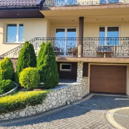 Jasna, dwukondygnacyjna elewacja domu z balkonem i ozdobną balustradą, otoczona zadbanym ogrodem z tujami i kamiennym murkiem, widok na podjazd z kostki brukowej.