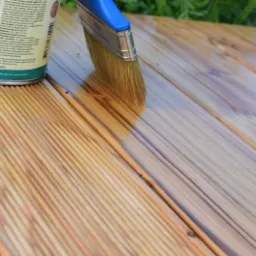 Lakierowanie desek tarasowych pędzlem, widoczna puszka z lakierem i świeżo nałożona warstwa ochronna.