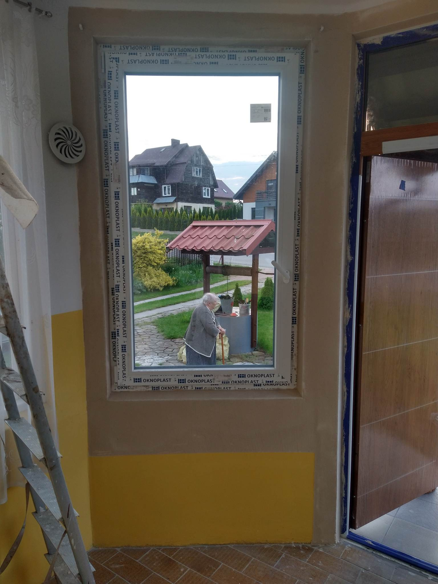 Nowe okno PCV marki Oknoplast, widok na ogród z studnią i starszą kobietą, w tle domy z ciemnego drewna, ściana wewnątrz w kolorach żółtym i beżowym, widoczna drabina.