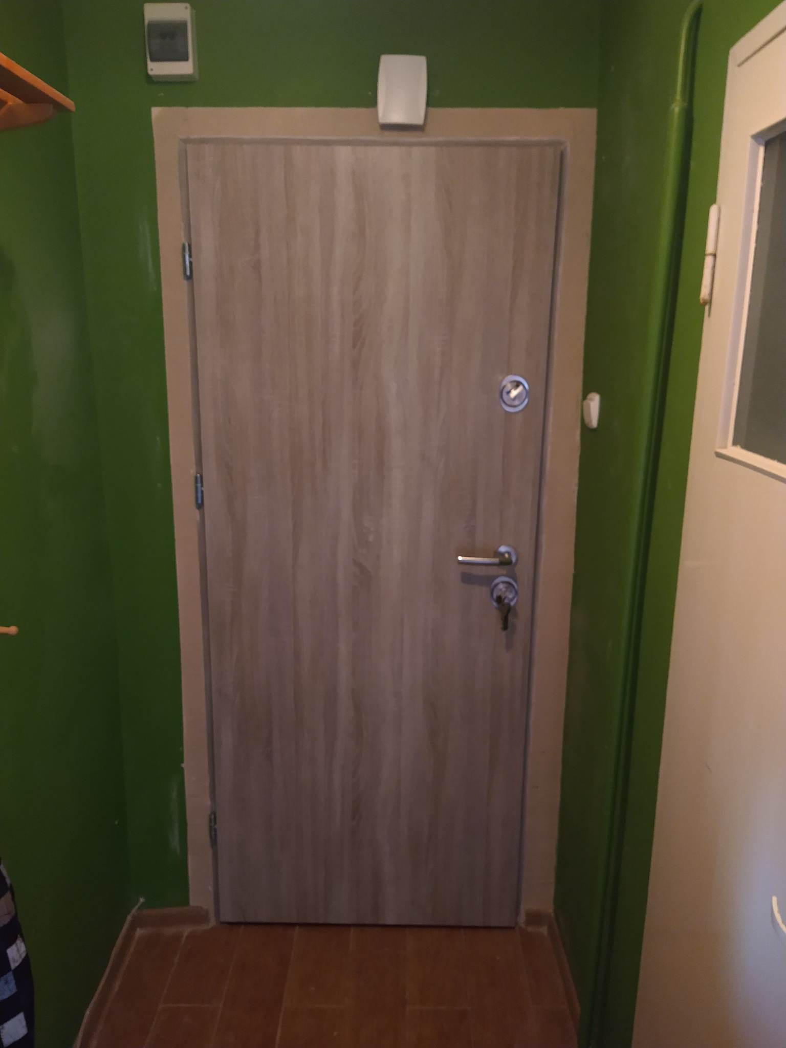 Zamknięte, drewnopodobne drzwi wejściowe z wizjerem i zamkiem z kluczem, osadzone w ościeżnicy w zielonej ścianie korytarza.