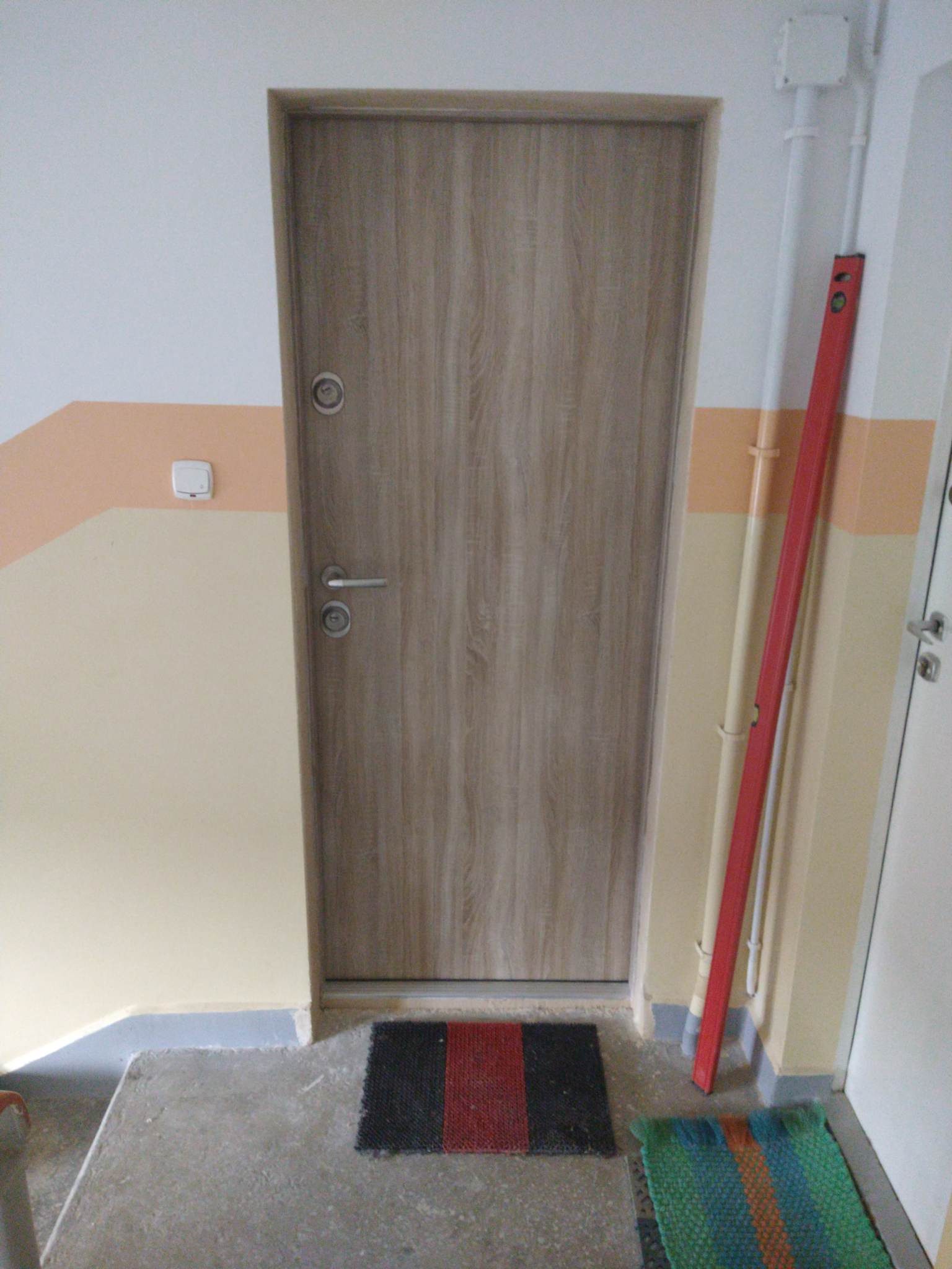 Drzwi wejściowe wewnątrz klatki schodowej, widoczny poziomica oparta o ścianę, dywaniki przed drzwiami.