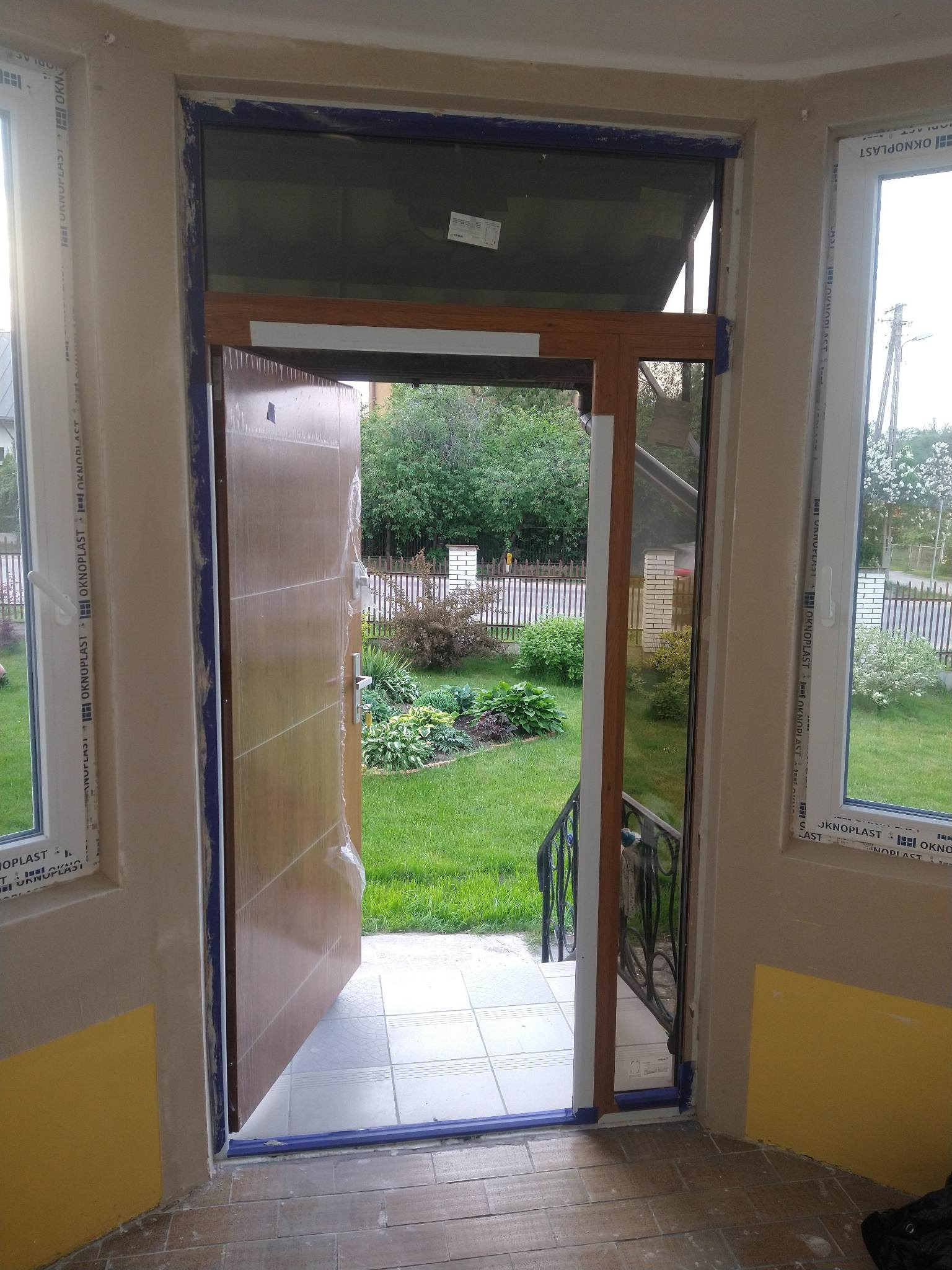Montaż drzwi balkonowych z folią ochronną, widoczne okna PCV z szybami, zabezpieczone taśmą malarską ściany, widok na ogród z zewnątrz