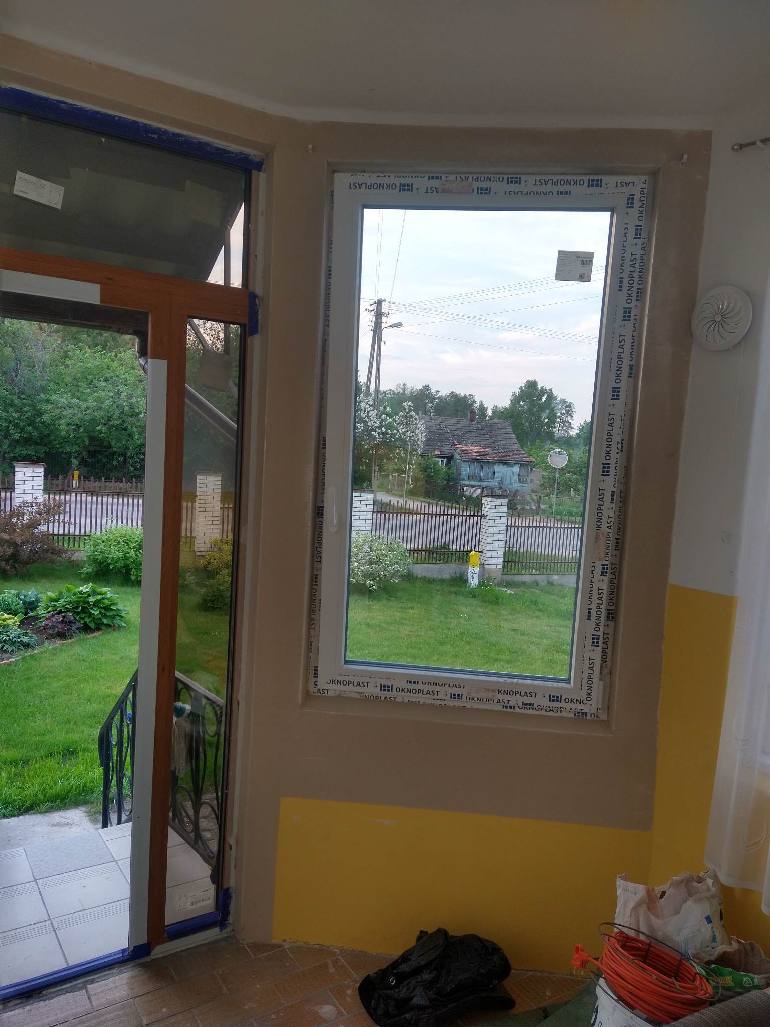 Montaż okna PCV marki Oknoplast z widokiem na ogród i dom jednorodzinny, widoczne ślady prac wykończeniowych na ścianie.