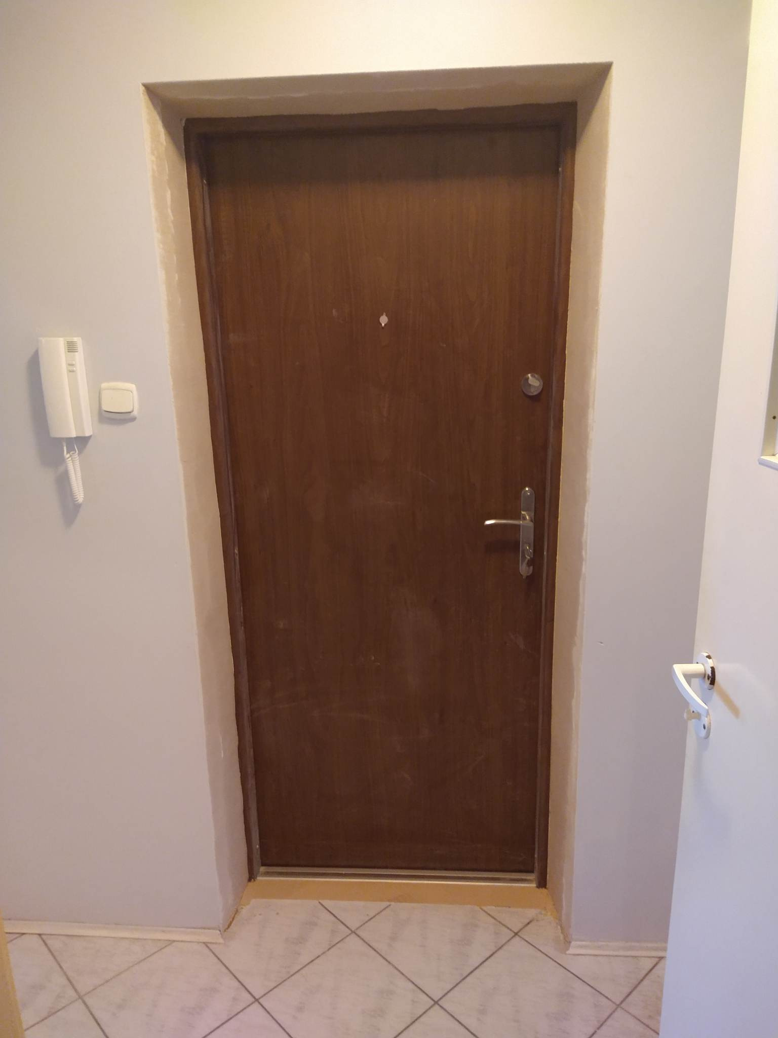 Brązowe drzwi wejściowe do mieszkania, widoczne ślady po malowaniu na ościeżnicy i ścianie, biały domofon po lewej stronie.