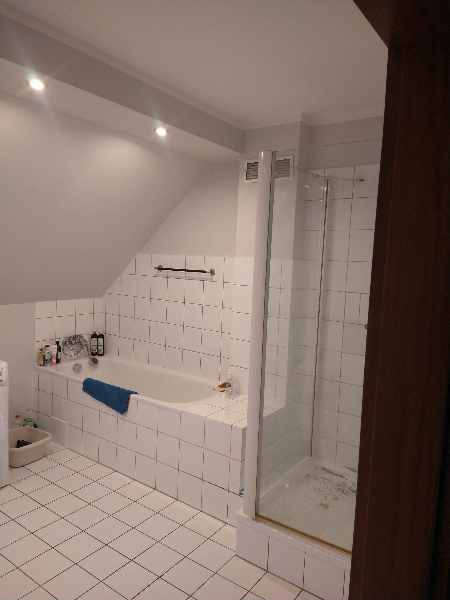 Biała łazienka na poddaszu z wanną obłożoną kafelkami, niebieskim ręcznikiem i kabiną prysznicową z widocznymi zaciekami na podłodze.