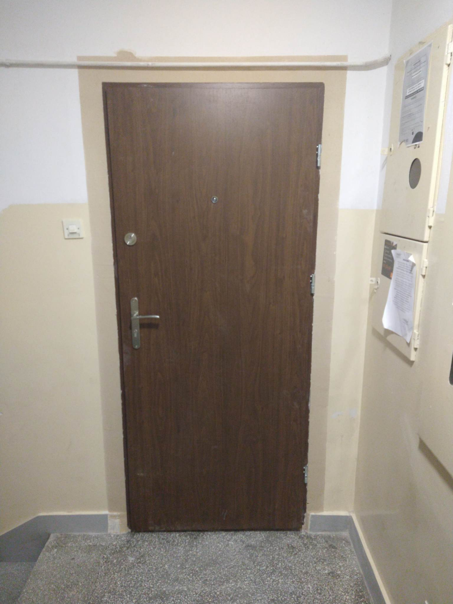 Brązowe drzwi wejściowe do mieszkania, widoczne z bliska na tle beżowych ścian i szarej podłogi, obok skrzynka elektryczna z dokumentami.