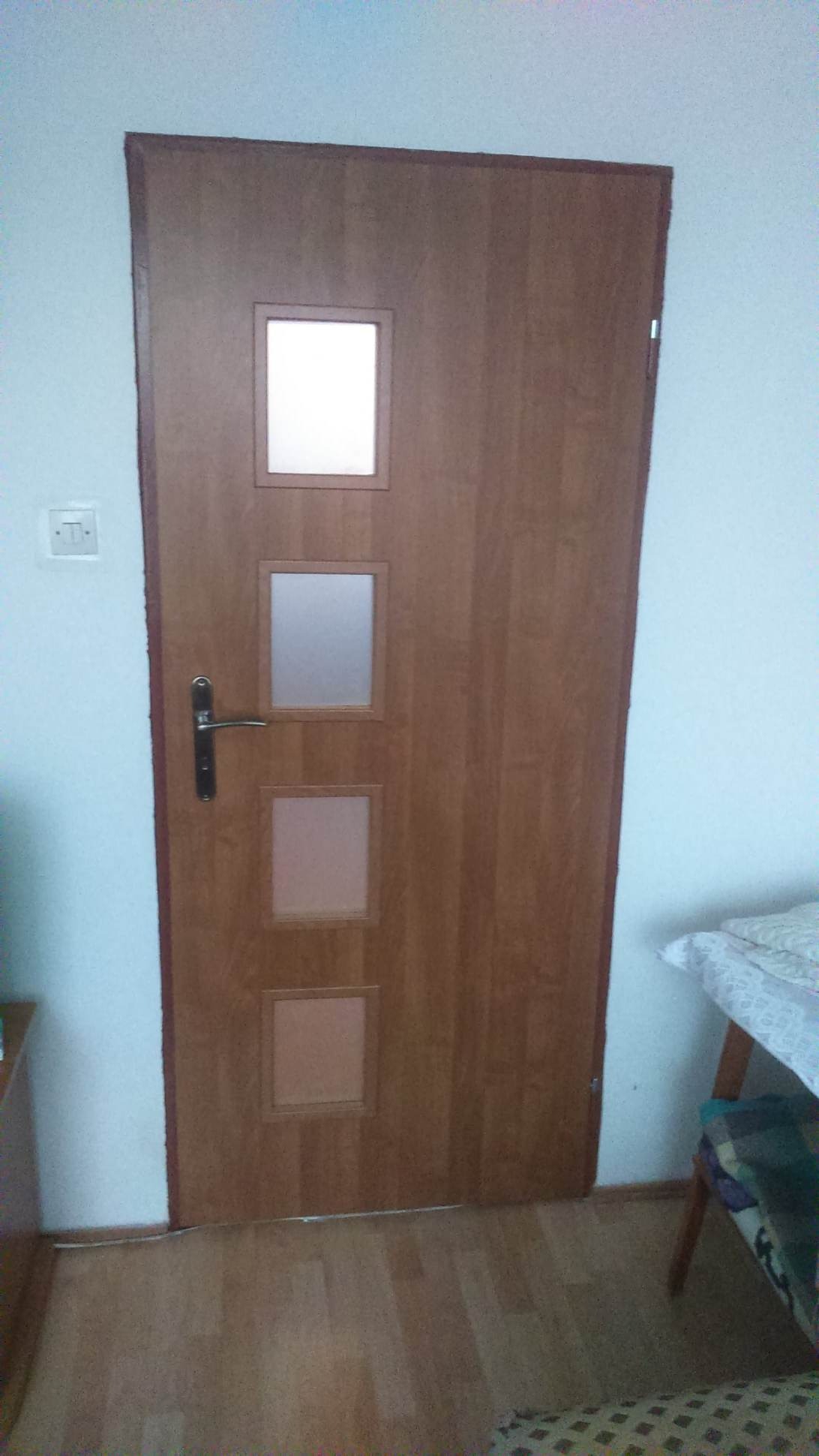 Drewniane drzwi wewnętrzne z czterema kwadratowymi przeszkleniami o różnym stopniu przezroczystości, zamontowane w białej ścianie, widoczne na tle drewnianej podłogi i fragmentu stołu z nakryciem.