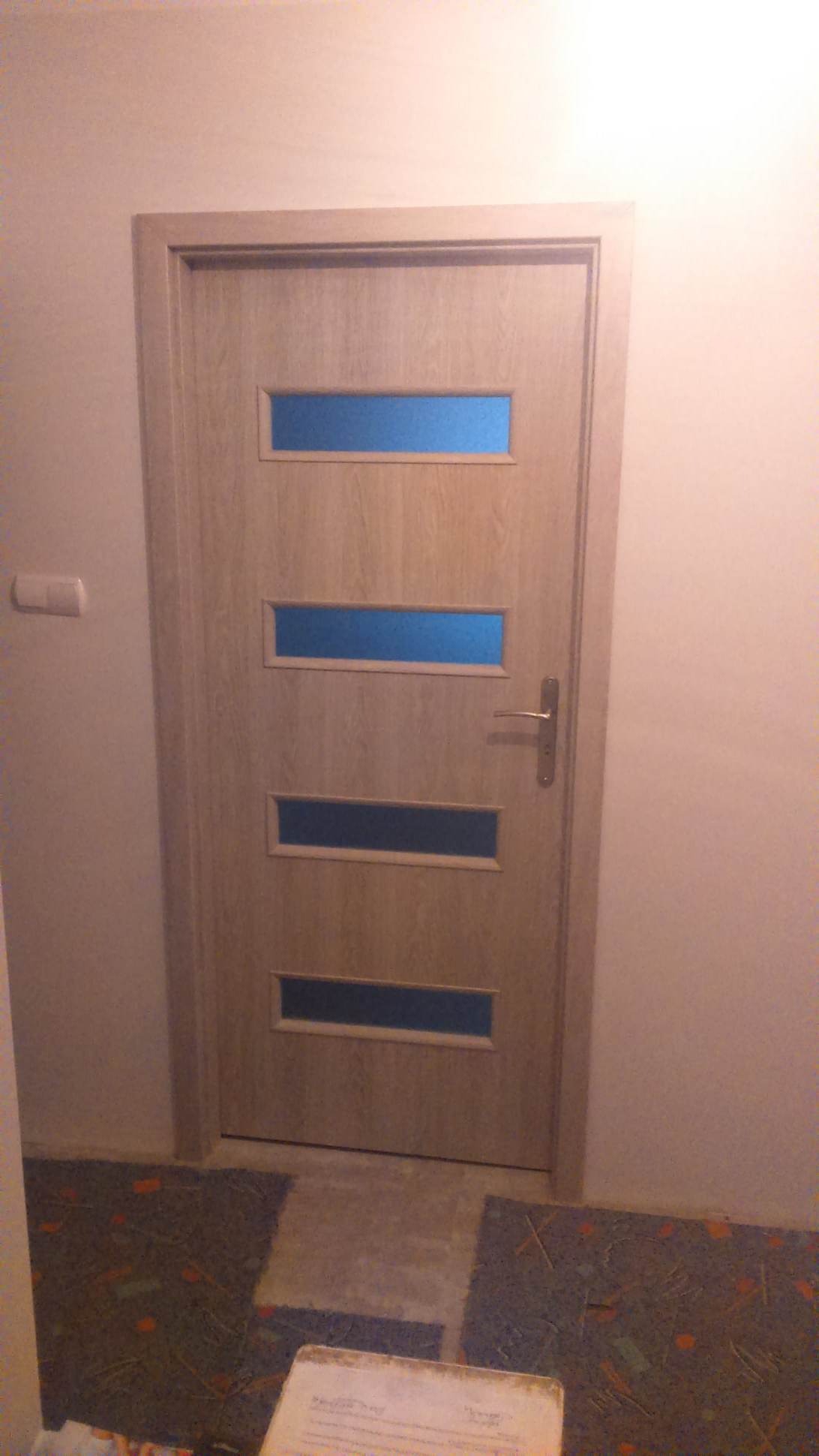Drewniane drzwi wewnętrzne z czterema poziomymi szybkami w kolorze niebieskim, zamontowane w mieszkaniu w Białymstoku. Widoczne ślady po remoncie na podłodze.