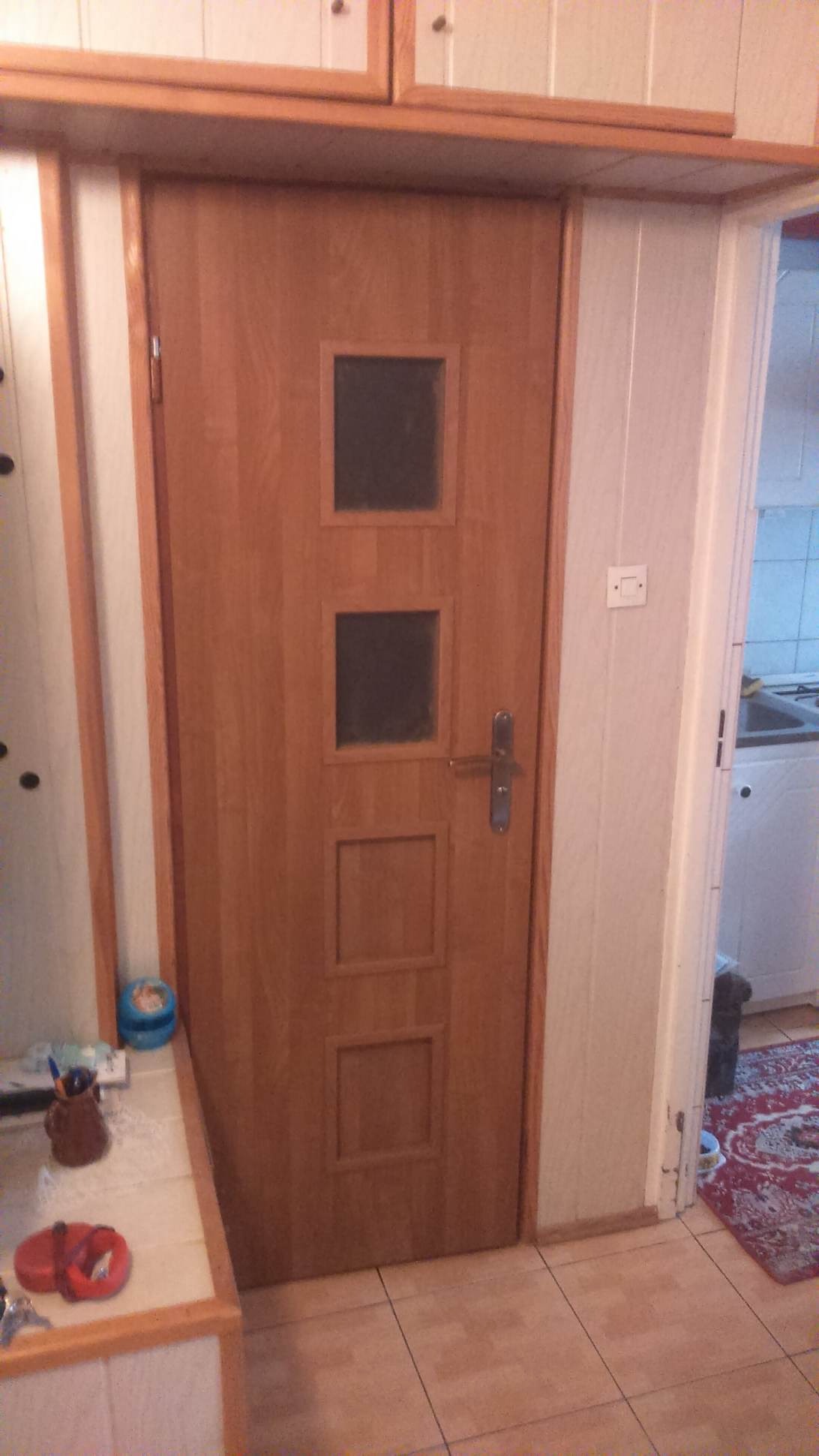 Drewniane drzwi wewnętrzne z trzema kwadratowymi przeszkleniami, zamontowane w ościeżnicy w pomieszczeniu z kafelkami na podłodze i widocznymi elementami wyposażenia kuchni w tle.