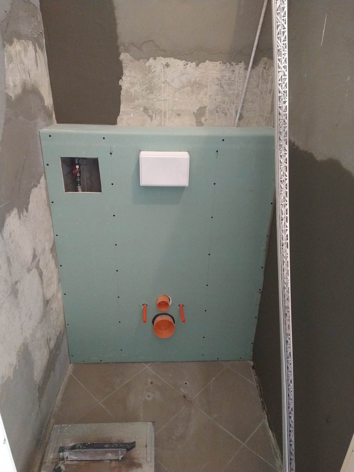 Niedokończona łazienka: ściana z płyty gipsowej z widocznymi rurami, spłuczka podtynkowa i nieotynkowane ściany, widoczna listwa narożna.