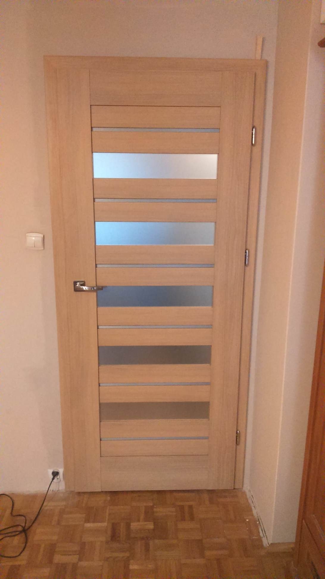 Jasne, drewniane drzwi wewnętrzne z poziomymi, matowymi szybami, zamontowane w ścianie. Widoczny parkiet na podłodze i gniazdko elektryczne z kablem.