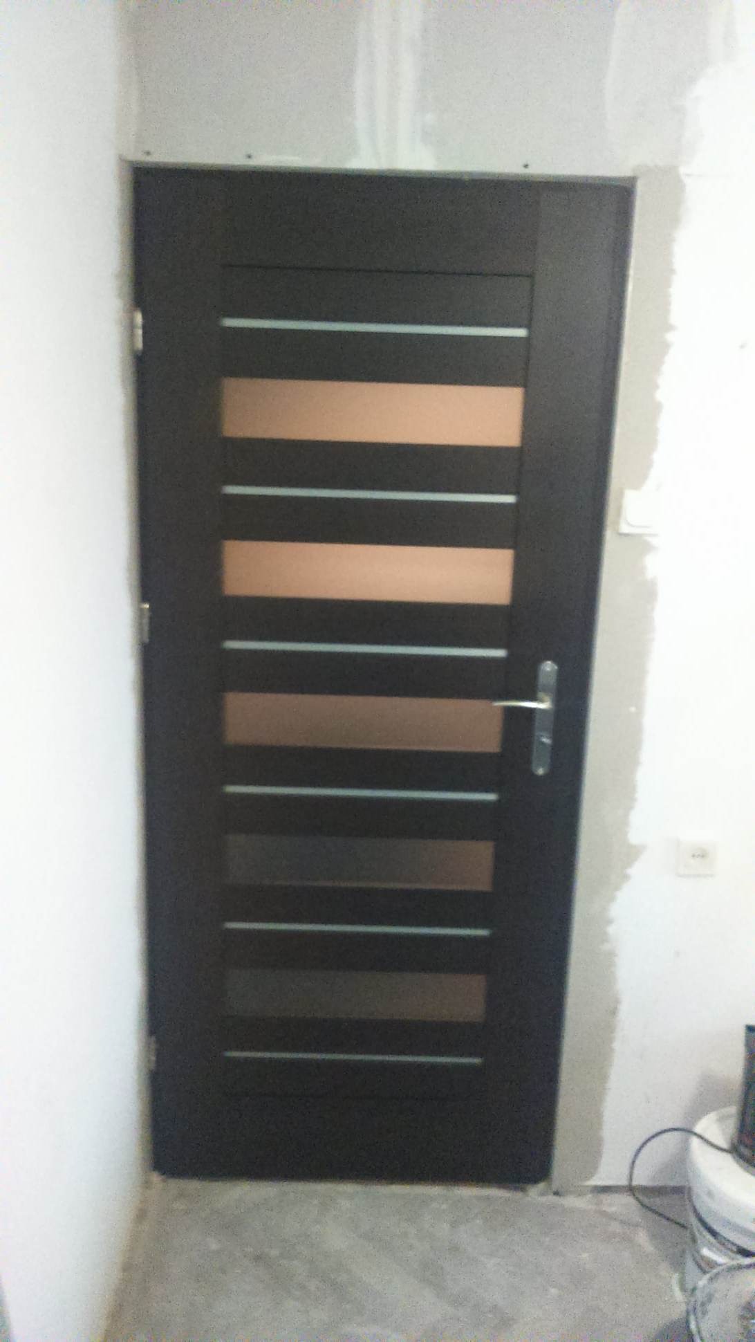 Ciemne drzwi wewnętrzne z poziomymi przeszkleniami, widoczne niedoskonałości wykończenia ściany i podłogi, sugerujące etap montażu.