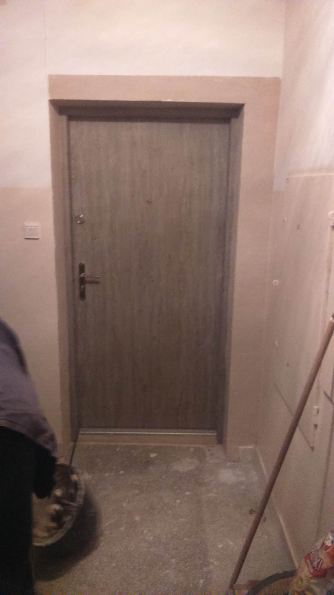 Montaż ościeżnicy i futryny wokół stalowych drzwi wejściowych w trakcie prac wykończeniowych; widoczny fragment pracownika z wiadrem i narzędzia.