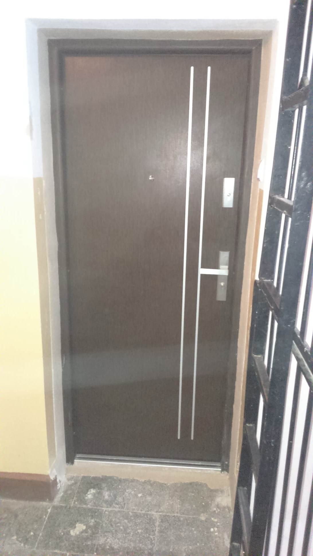 Brązowe drzwi wejściowe z dwoma pionowymi, srebrnymi paskami, zamontowane w bloku mieszkalnym, widoczne niedoskonałości wykończenia przy podłodze.