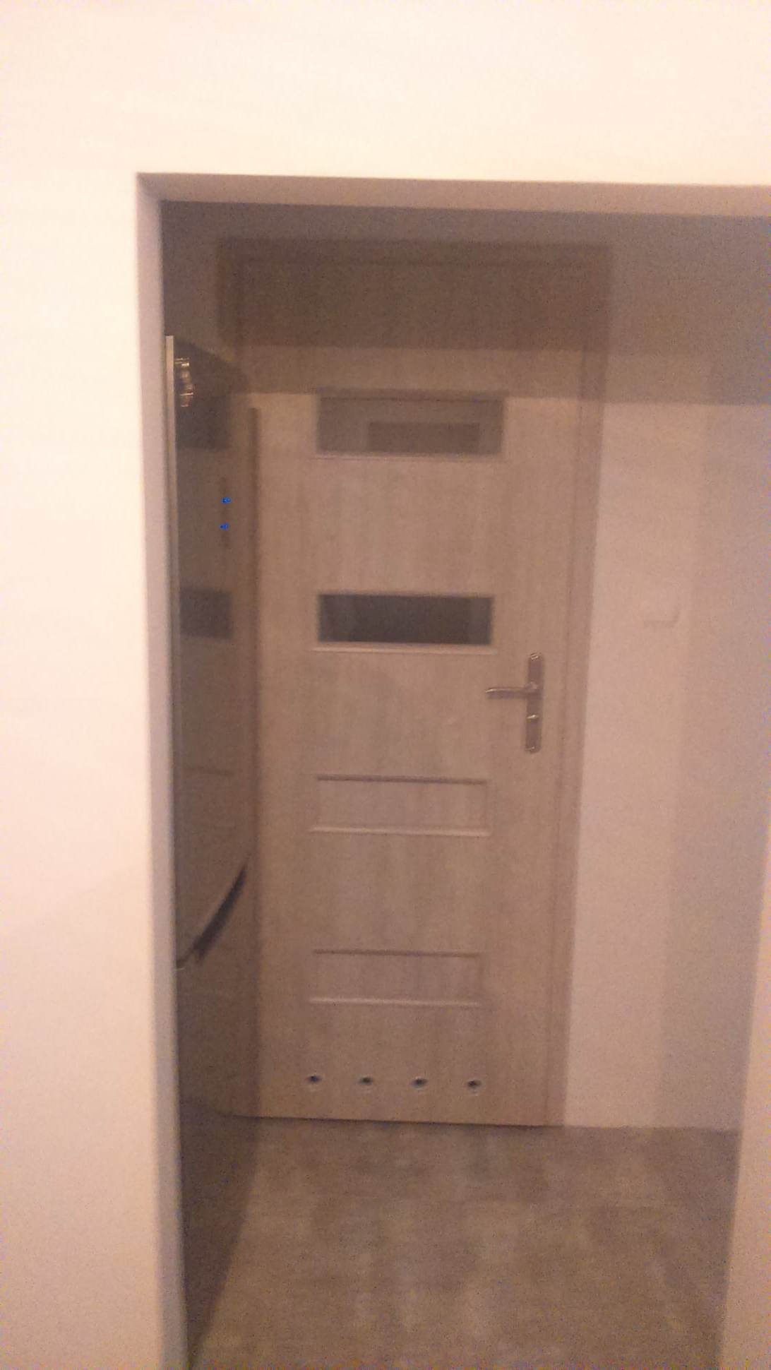 Nowe, jasne drzwi wewnętrzne z przeszkleniem, zamontowane w świeżo pomalowanym pomieszczeniu. Widoczne otwory montażowe w dolnej części.