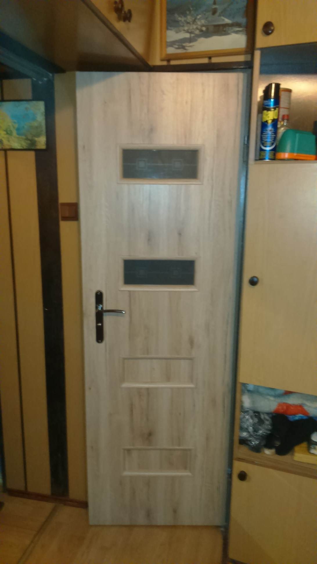 Zamknięte drzwi wewnętrzne z drewnopodobnym wzorem i dwoma przeszkleniami, widoczne w pomieszczeniu z meblami.