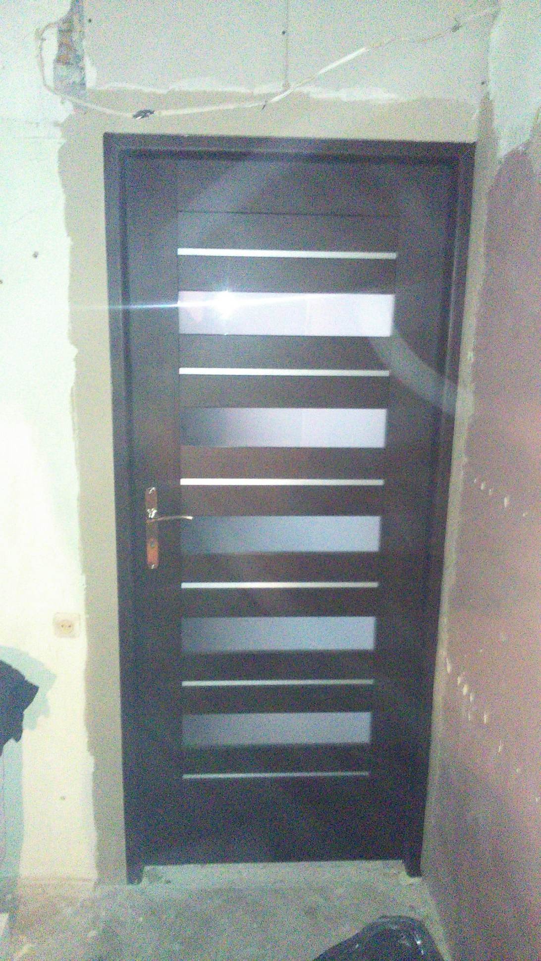 Ciemne, nowoczesne drzwi wewnętrzne z poziomymi, prostokątnymi przeszkleniami, zamontowane w surowym otworze w ścianie, widoczne ślady prac wykończeniowych na ścianach i podłodze.