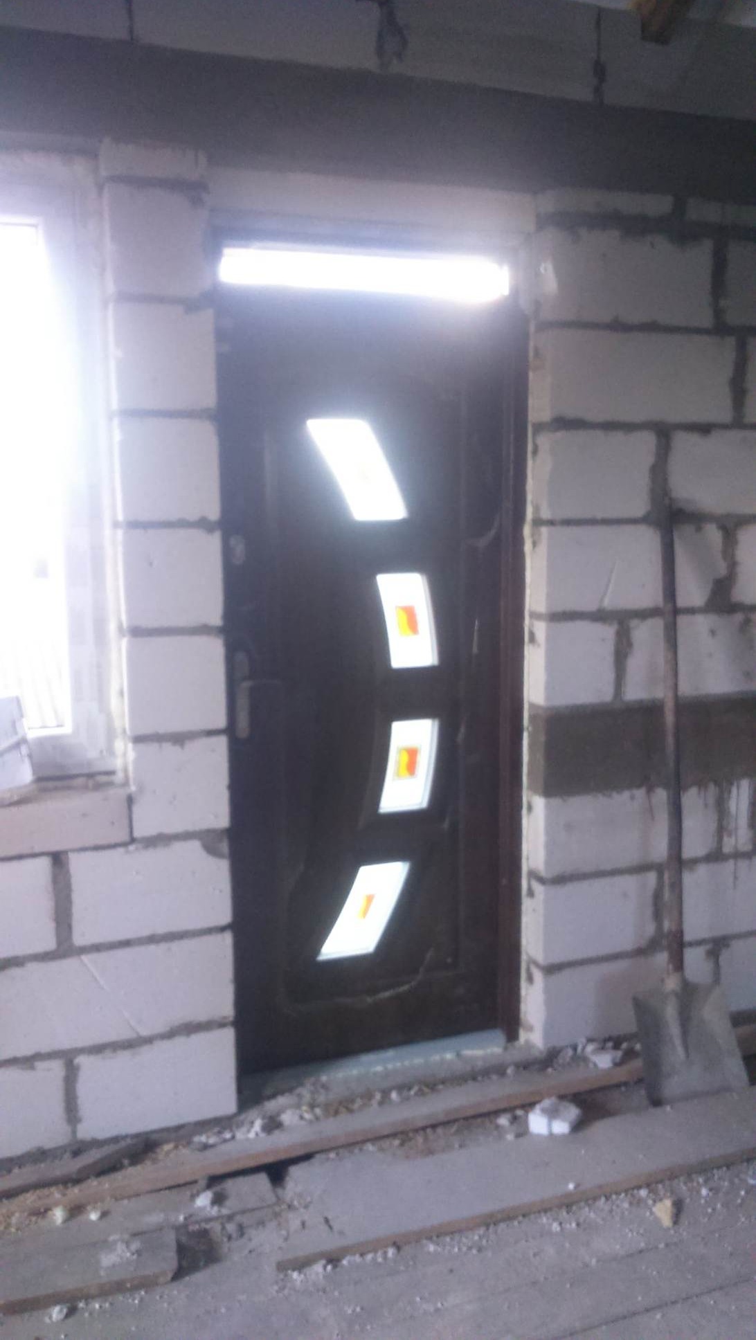 Montaż brązowych drzwi wejściowych z trzema podświetlanymi, pionowo ułożonymi prostokątnymi szybkami z pomarańczowymi akcentami w surowym otworze murowanym z białych bloczków betonowych. Widoczna...