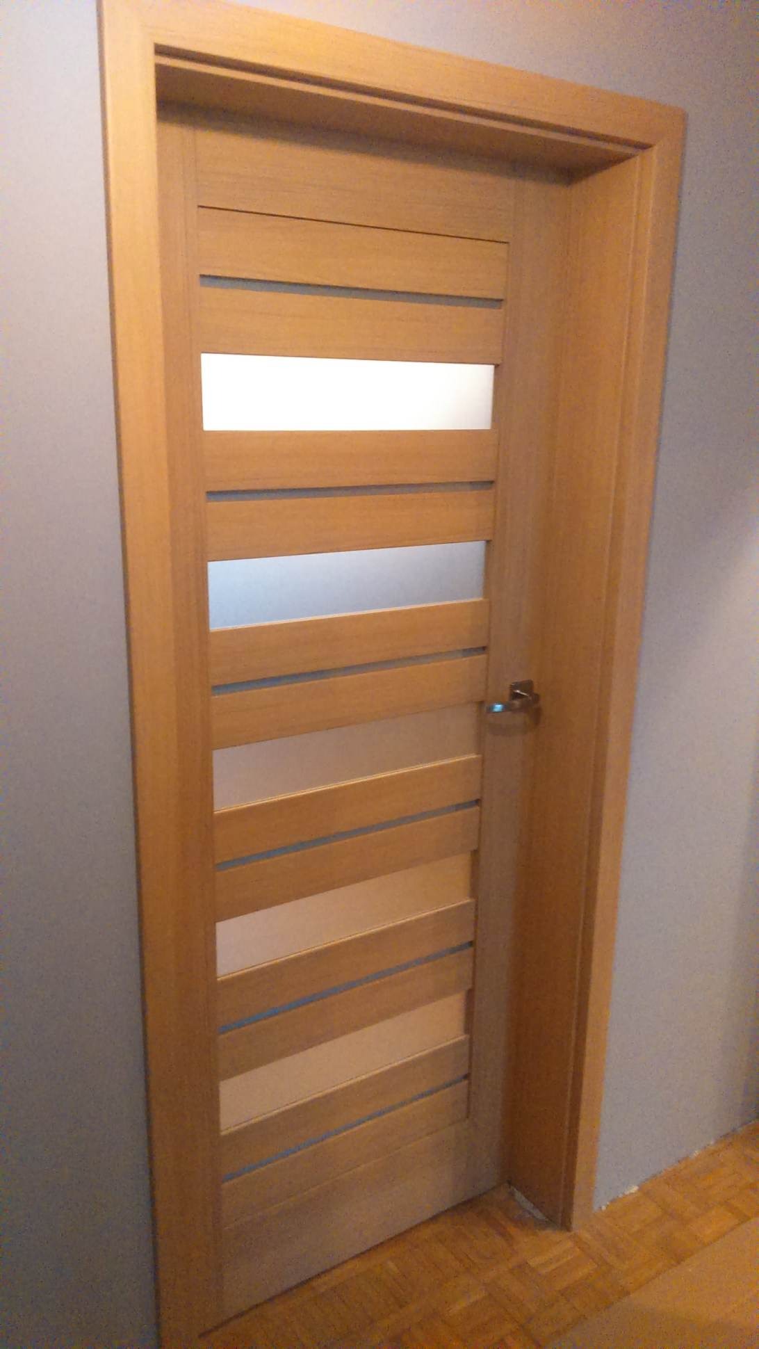 Zamknięte, drewniane drzwi wewnętrzne z poziomymi, przeszklonymi paskami, widoczne na tle szarej ściany i parkietu.