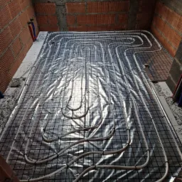 Montaż ogrzewania podłogowego: białe rurki ułożone na czarnej siatce, pokrytej folią termoizolacyjną, widoczne fragmenty ceglanej ściany i sufitu w surowym stanie.