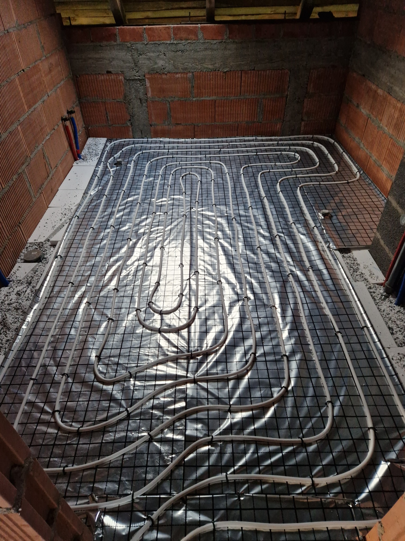 Montaż ogrzewania podłogowego: białe rurki ułożone na czarnej siatce, pokrytej folią termoizolacyjną, widoczne fragmenty ceglanej ściany i sufitu w surowym stanie.