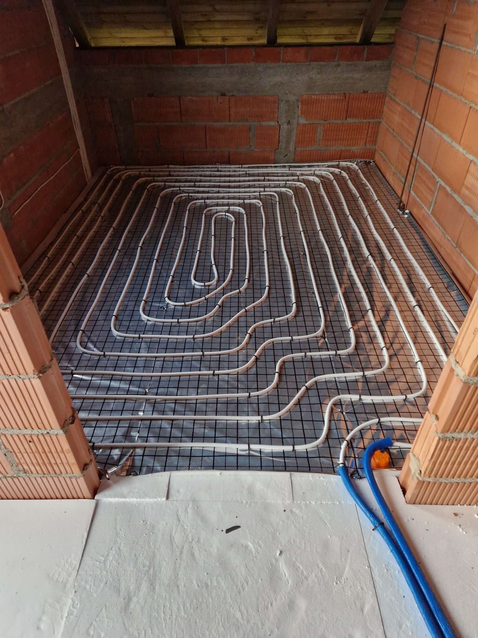 Montaż ogrzewania podłogowego: białe rurki ułożone w pętle na czarnej siatce, widoczne fragmenty ceglanej ściany i izolacji podłogowej, dwa niebieskie przewody doprowadzające.