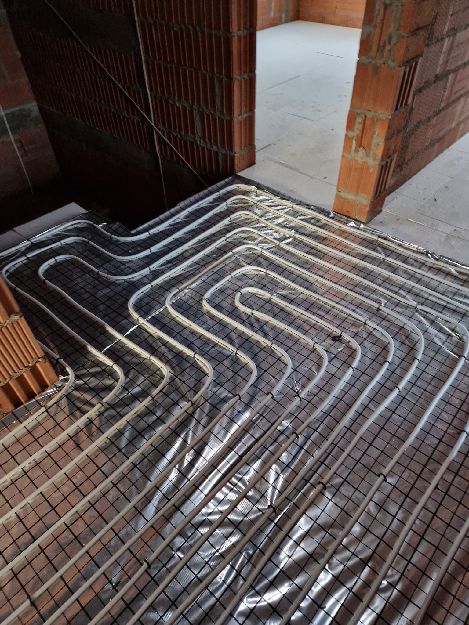 Białe rury ogrzewania podłogowego ułożone na srebrnej folii izolacyjnej z czarną siatką, widoczne surowe ściany z czerwonej cegły w nowym budynku.